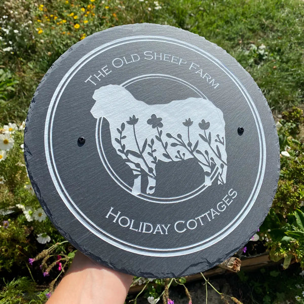 Round slate sign with sheep design and text 'The Old Sheep Farm Holiday Cottages' held in front of a grassy background.