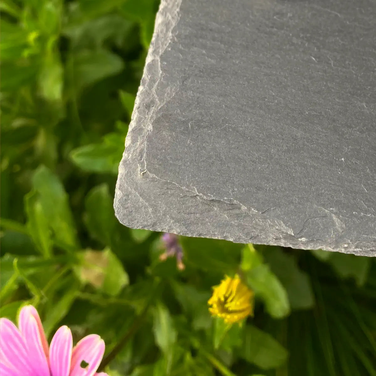 “Close-up of a chiseled edge on a Welsh slate sign with a natural, textured finish, shown against a background of green foliage and colorful flowers.”