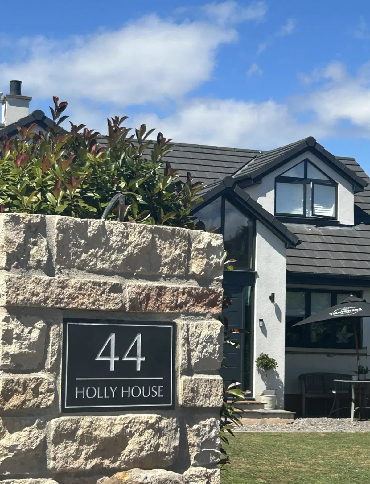 A black rectangular house number plaque reading '44 HOLLY HOUSE' mounted on a rough stone wall, with part of a modern house and outdoor patio visible in the background.