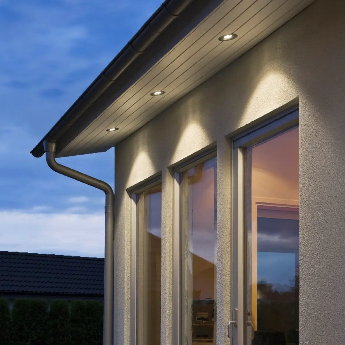 An exterior view of a modern house at dusk, featuring three large windows and an overhanging roof with recessed downlights. The lights cast warm, focused beams onto the wall below. The sky is a deepening blue, and the interior of the house is softly illuminated, visible through the windows. A metal gutter runs along the edge of the roof.