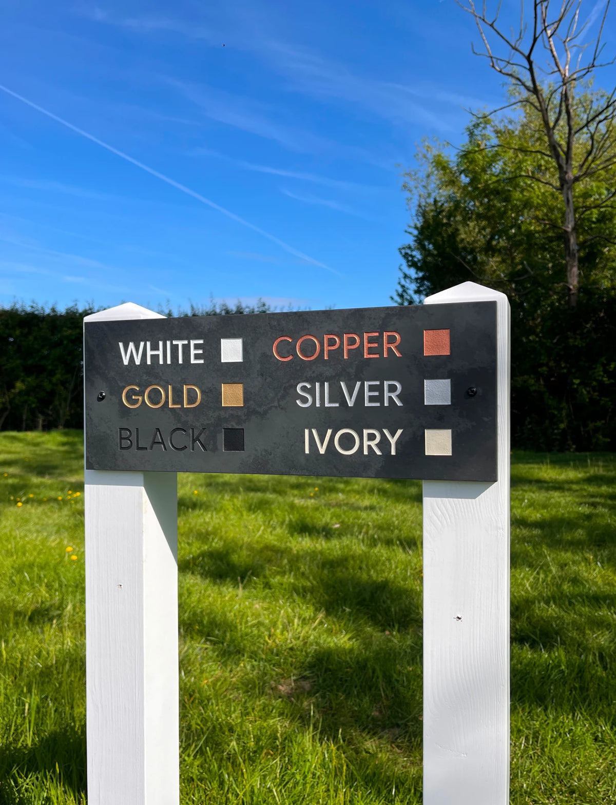 A black metal sign on white posts displaying color options for a product, with white, copper, gold, silver, black, and ivory color blocks and labels, set against a grassy lawn and trees under a blue sky.