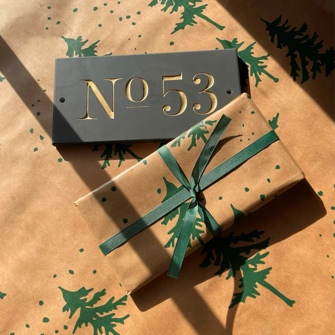 A slate house sign with the number “53” engraved in gold sits next to a neatly wrapped gift, placed on festive wrapping paper with green tree patterns.