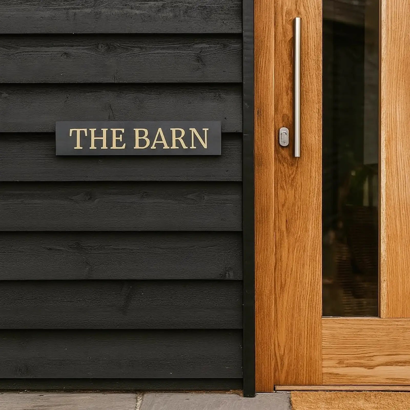 Metal sign with 'THE BARN' on a black weatherboard barn wall next to an oak door
