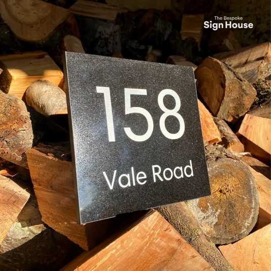 a black granite house sign with 158 vale road engraved and painted white. the sign is balancing on cut up logs