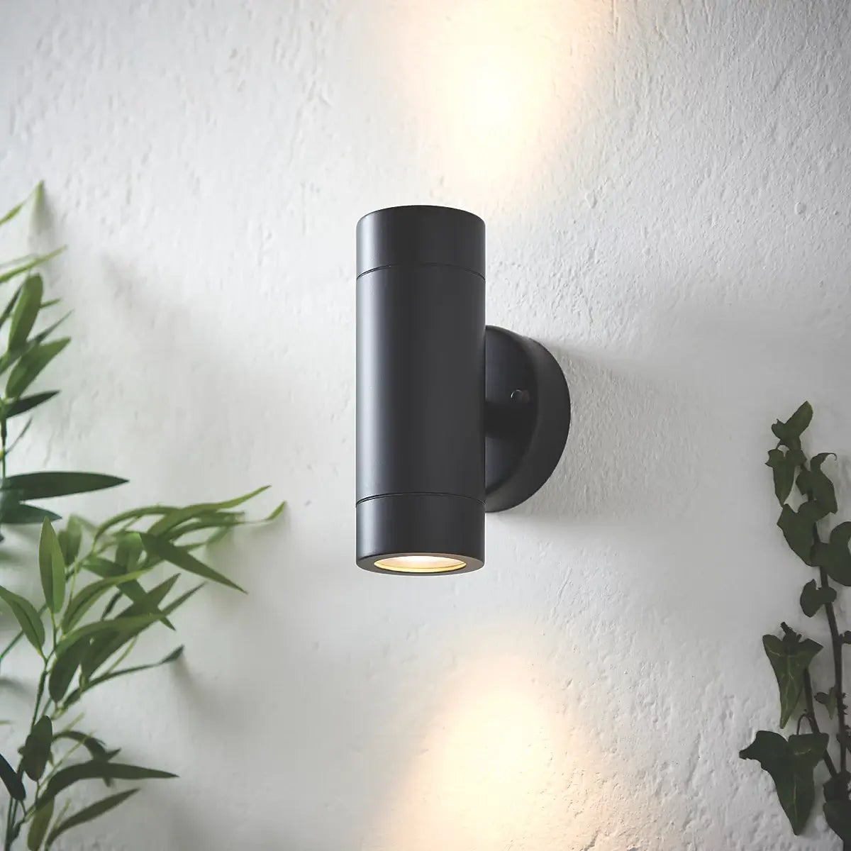 The image shows a modern grey cylindrical outdoor wall light mounted on a white textured wall. It emits both upward and downward light, creating a stylish and balanced illumination. Greenery on either side enhances the sleek and contemporary look of the fixture.