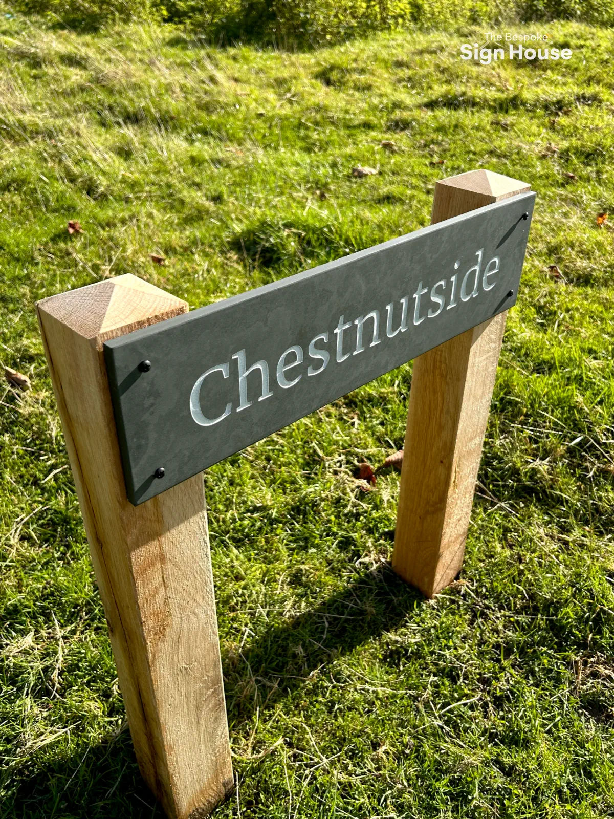 The image shows a grey slate house sign mounted on sturdy oak posts with pyramid tops, set in a grassy area. The sign displays the word “Chestnutside” in white, engraved lettering. The natural tones of the wood and slate contrast nicely with the vibrant green grass, creating a stylish and durable outdoor display. The Bespoke Sign House logo is visible in the upper right corner.