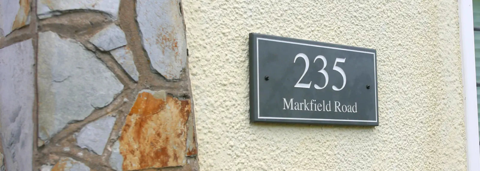 A banner featuring a variety of house signs, including wall-mounted and freestanding designs, crafted in slate with elegant text. The signs are displayed on different backdrops, such as brick, wood, stone, and painted walls. A man is shown smiling and pointing at one of the signs.