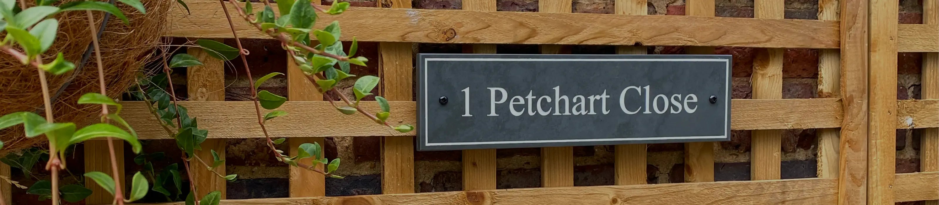 A banner featuring a variety of house signs, including wall-mounted and freestanding designs, crafted in slate with elegant text. The signs are displayed on different backdrops, such as brick, wood, stone, and painted walls. A man is shown smiling and pointing at one of the signs.