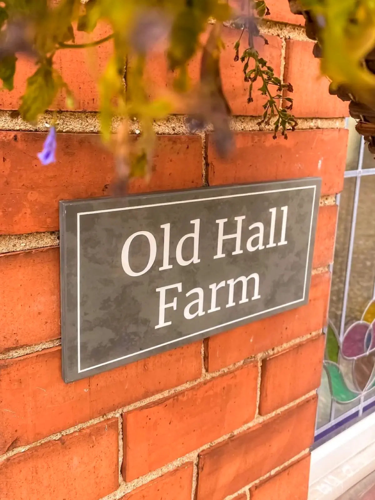 Close-up view of a custom house sign reading "Old Hall Farm," mounted on a red brick wall with some blurred greenery in the foreground.