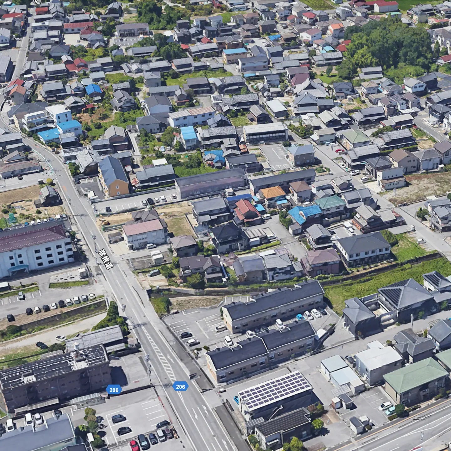 “Aerial view of a suburban area in Japan, featuring residential buildings, narrow streets, and parking areas, with a main road labeled Route 206 running through the scene.”