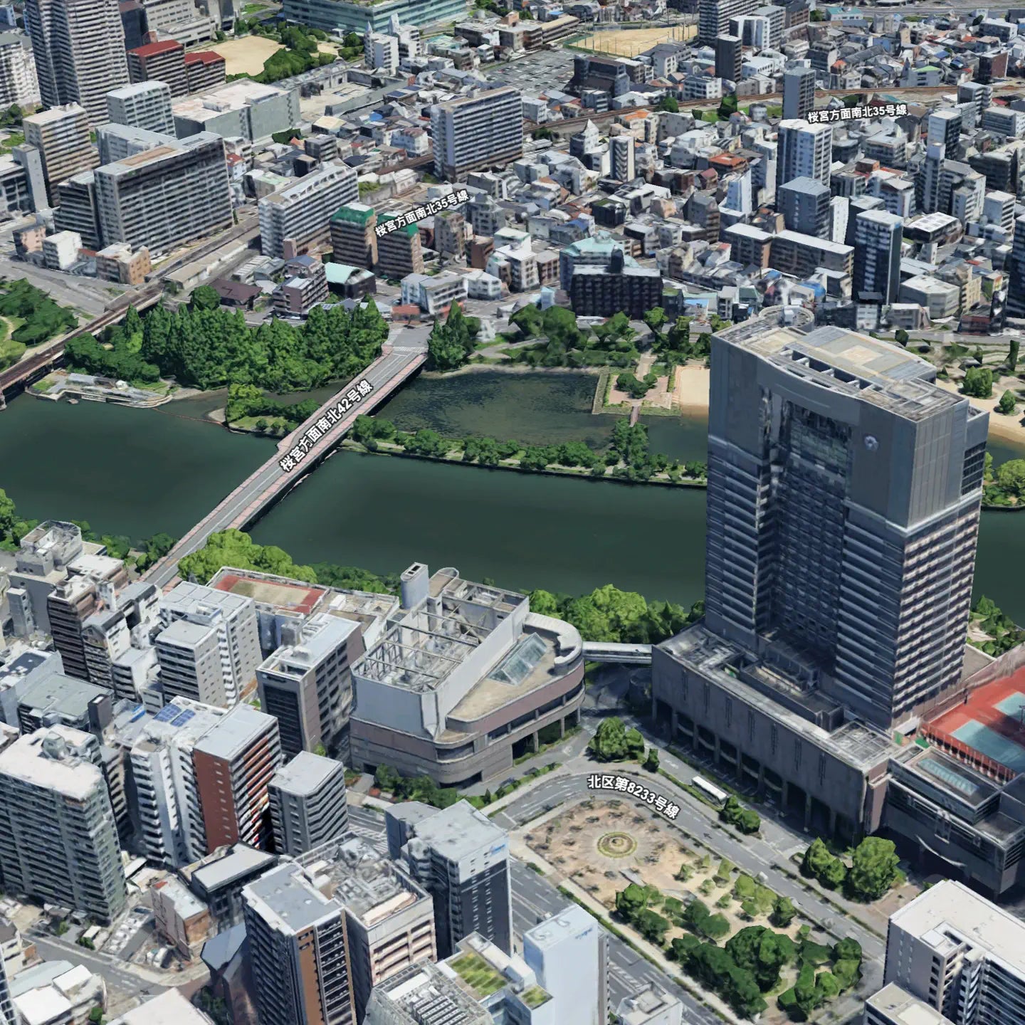 “Aerial view of an urban area in Japan, featuring a river, bridge, and high-rise buildings surrounded by densely packed city structures and green spaces.”