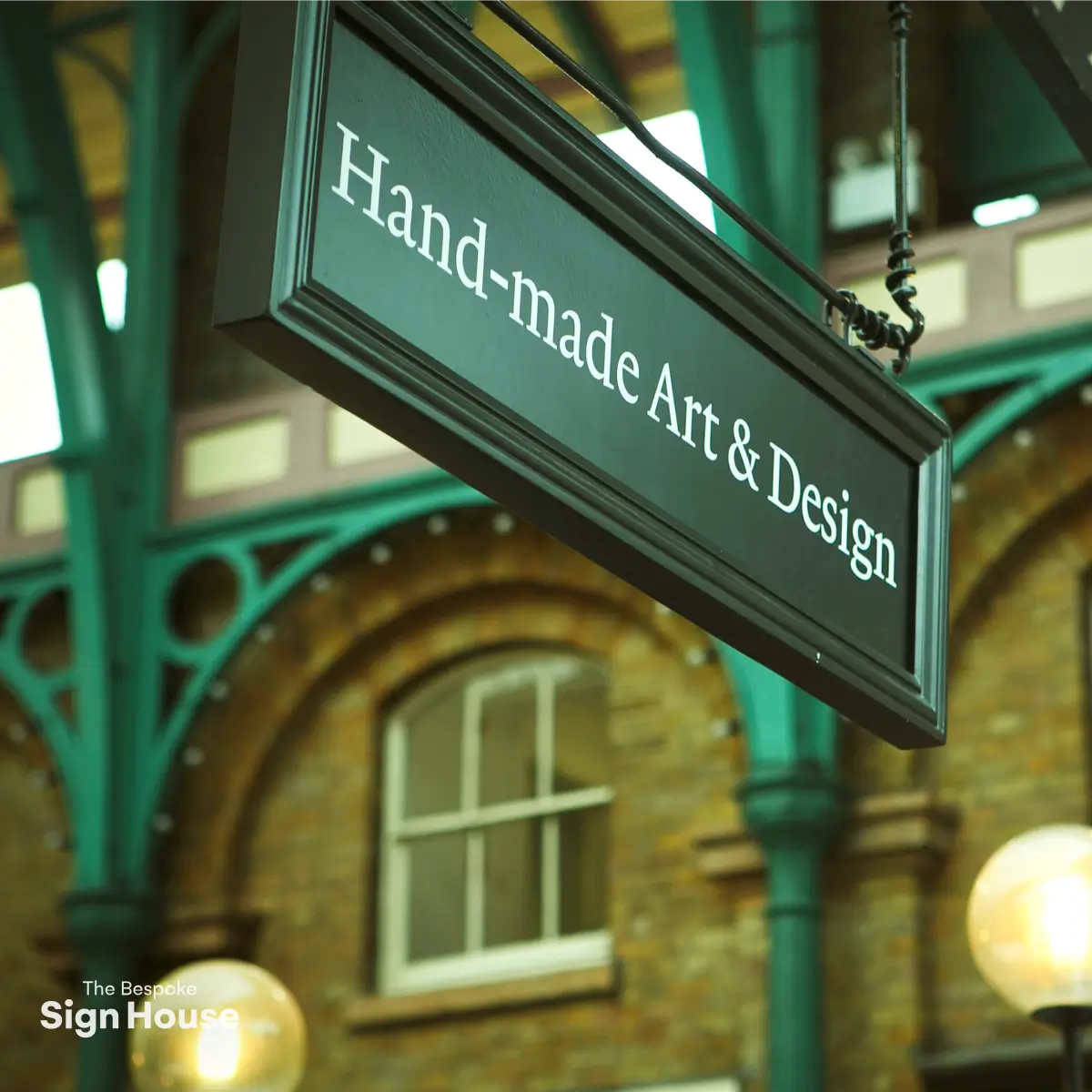 A black hanging sign reads "Hand-made Art & Design" against a background of green beams and brick architecture.