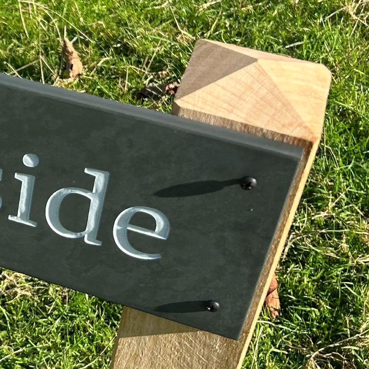 Close-up of a slate house sign with engraved lettering, securely mounted on an oak post with a pyramid top, against a grassy outdoor background.