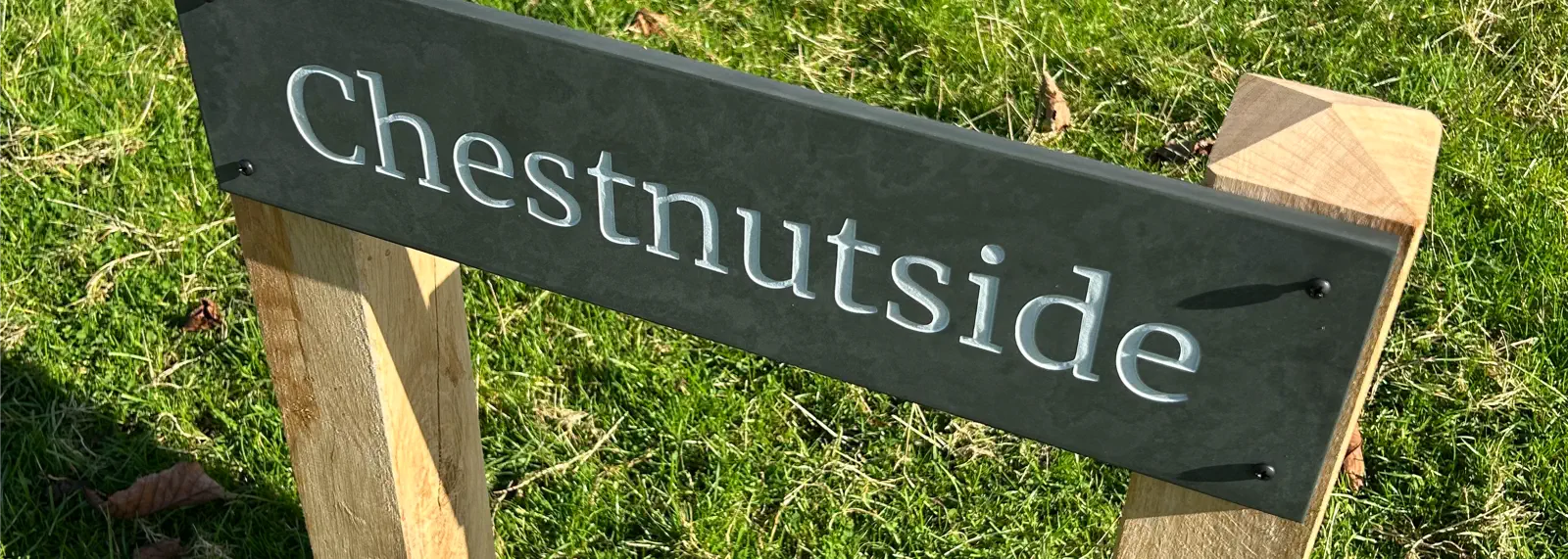 A banner showcasing two freestanding slate house signs mounted on sturdy oak posts, displayed in a lush green field. The left sign reads “Chestnutside” in elegant white text, while the right sign features “Ardoch House.” Both designs highlight the timeless combination of natural wood and slate, perfect for rural or garden settings.