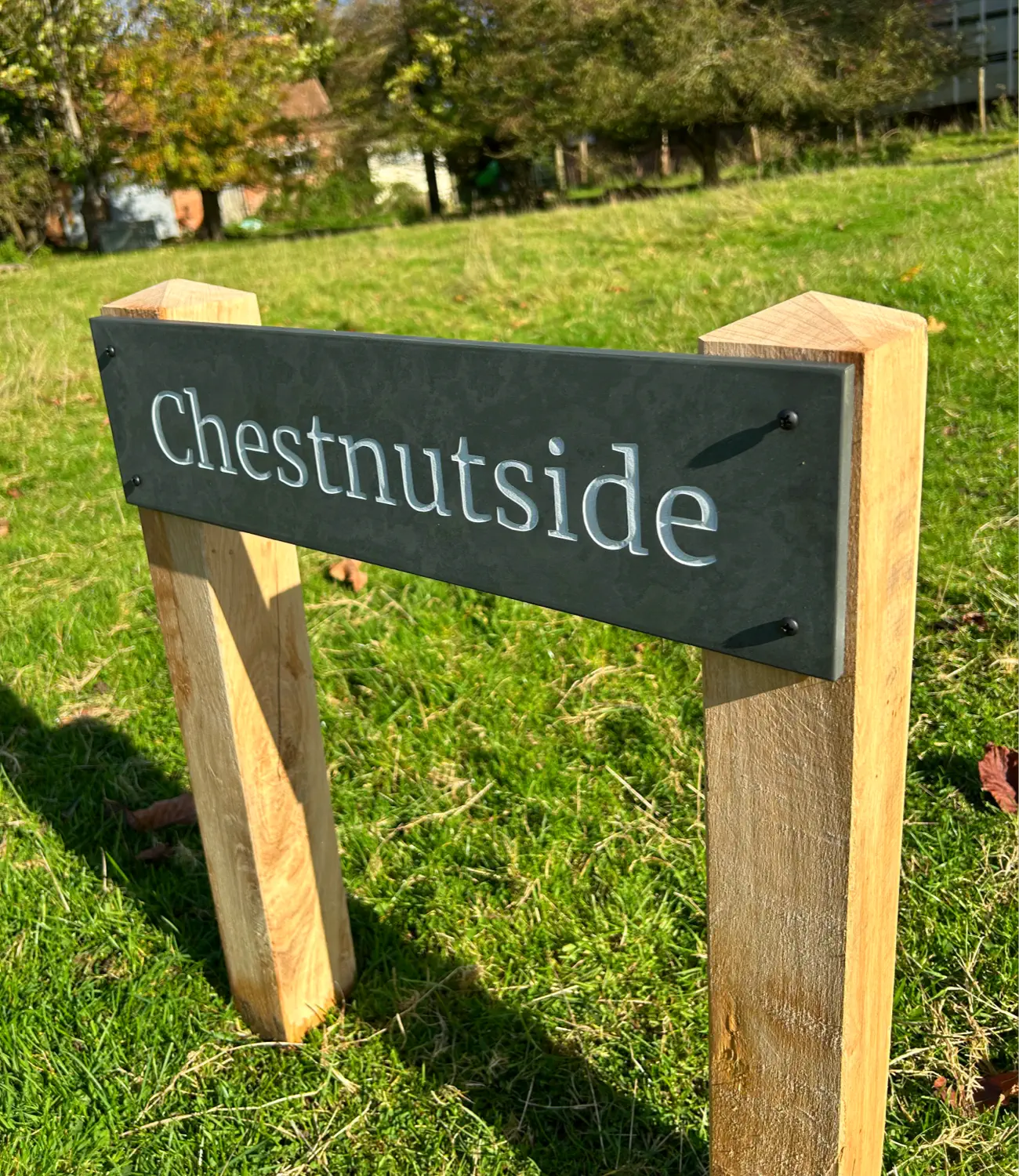 A close-up view of a freestanding slate house sign reading “Chestnutside,” securely mounted on oak posts. Set against a vibrant grassy backdrop, this sign demonstrates a stylish yet practical solution for marking entrances or property boundaries.