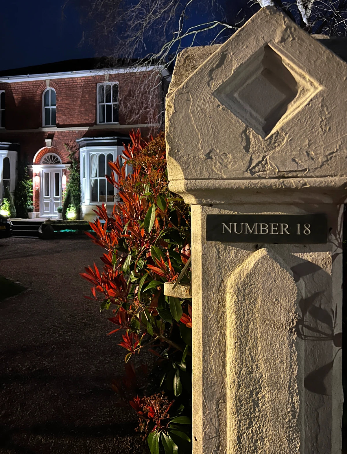A close-up of a beige stone pillar at the entrance of a house at night, with a black plaque reading 'NUMBER 18'. Red and green foliage is visible beside the pillar, and a brick building with lit windows appears in the background.