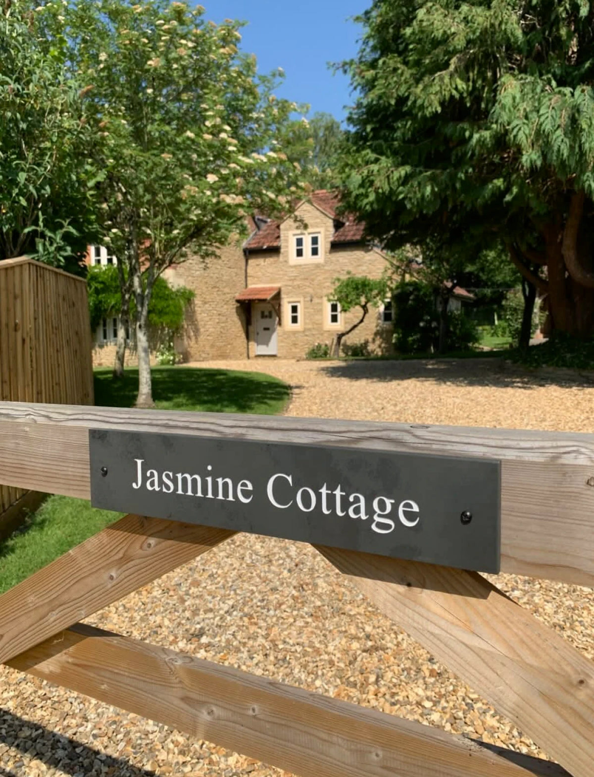 A custom black metal sign with white text reading 'Jasmine Cottage' mounted on a wooden gate in an outdoor setting with gravel and a house in the background.