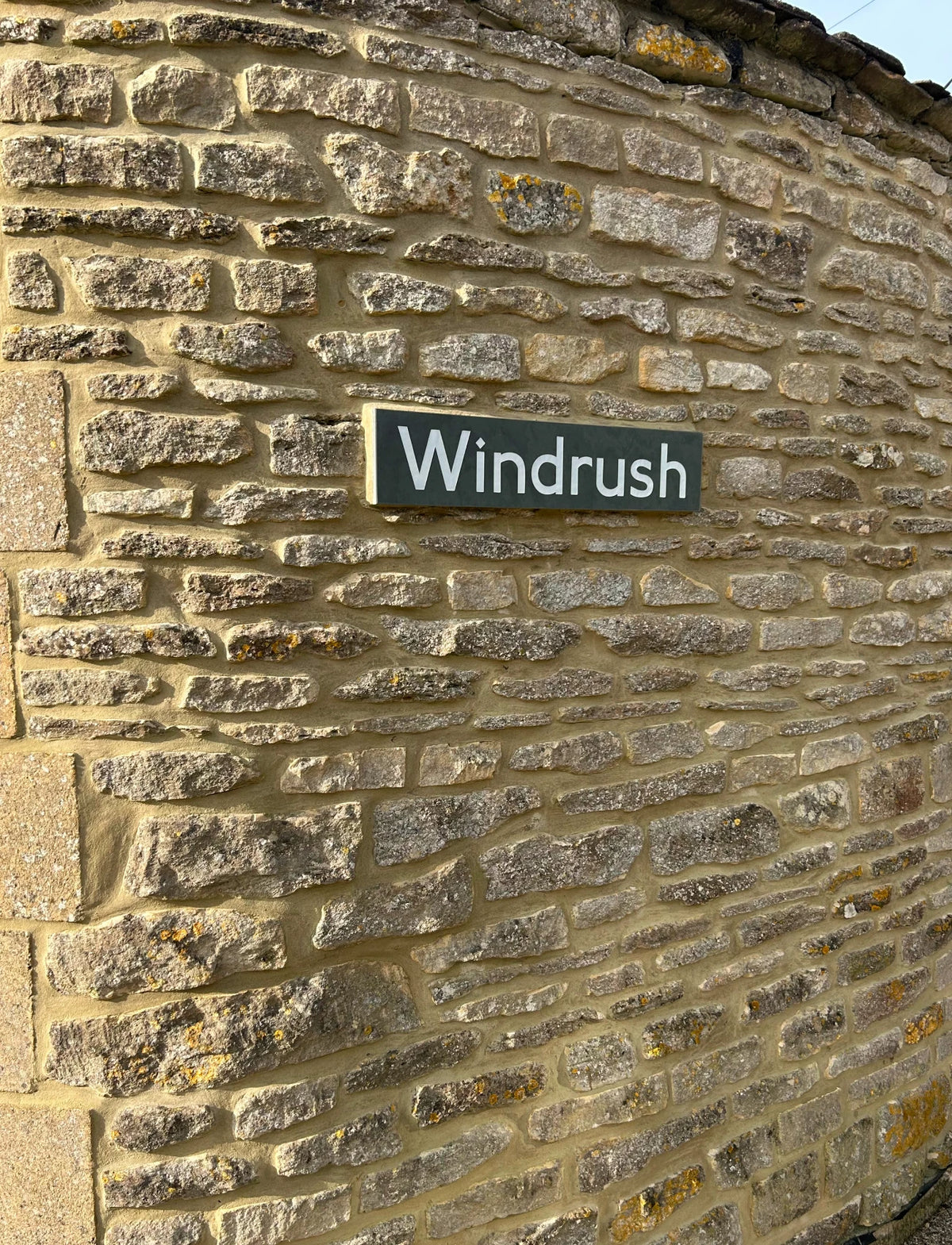 A rectangular black sign with the word 'Windrush' in white lettering, mounted on a textured stone wall.