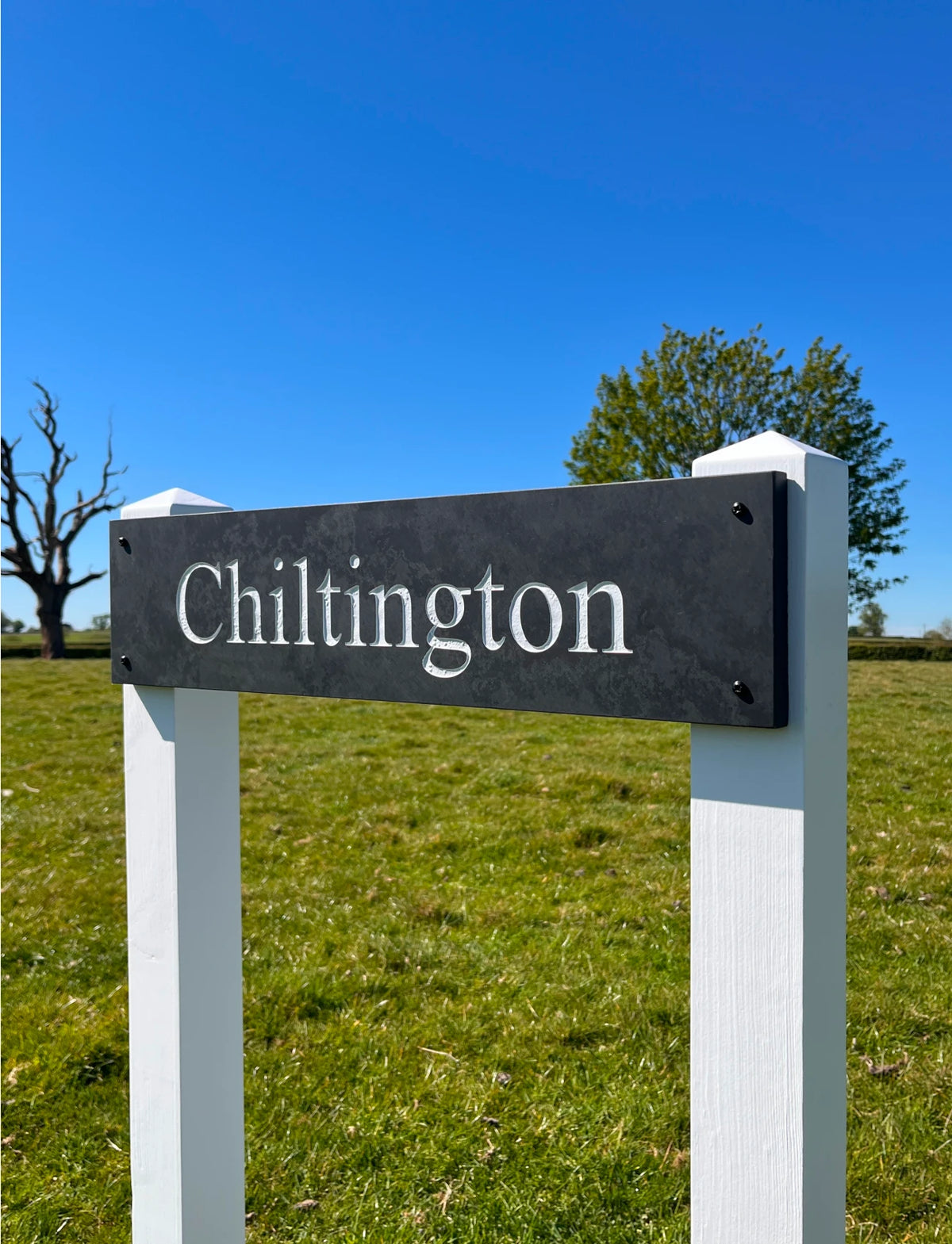 A black rectangular name sign with the word "Chiltington" in white serif letters, mounted on two white posts in a grassy field under a blue sky.
