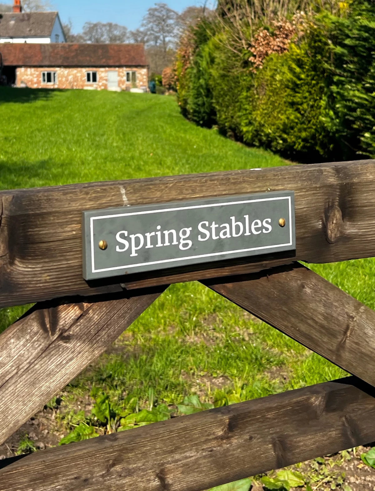 A rectangular metal sign reading 'Spring Stables' mounted on a weathered wooden gate, with a green lawn, trees, and a stone building in the background.
