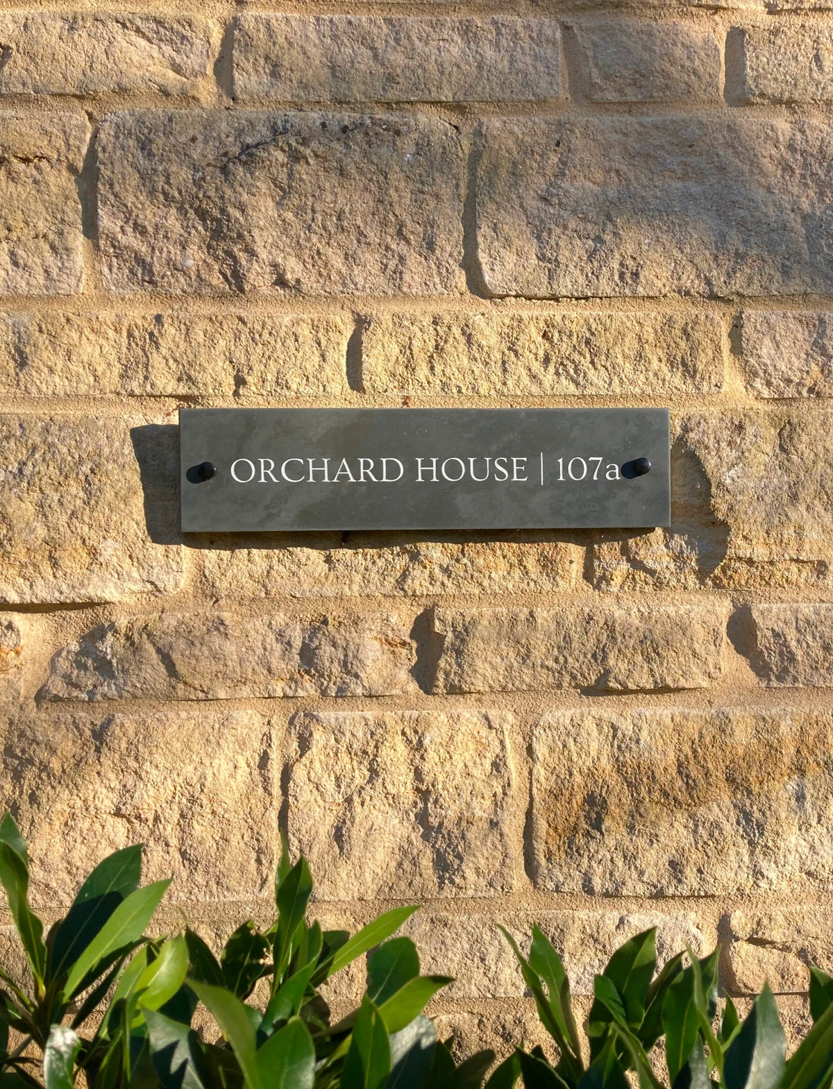 A dark rectangular metal house plaque reading 'ORCHARD HOUSE | 107a' mounted on a beige stone wall next to a dark wooden door.