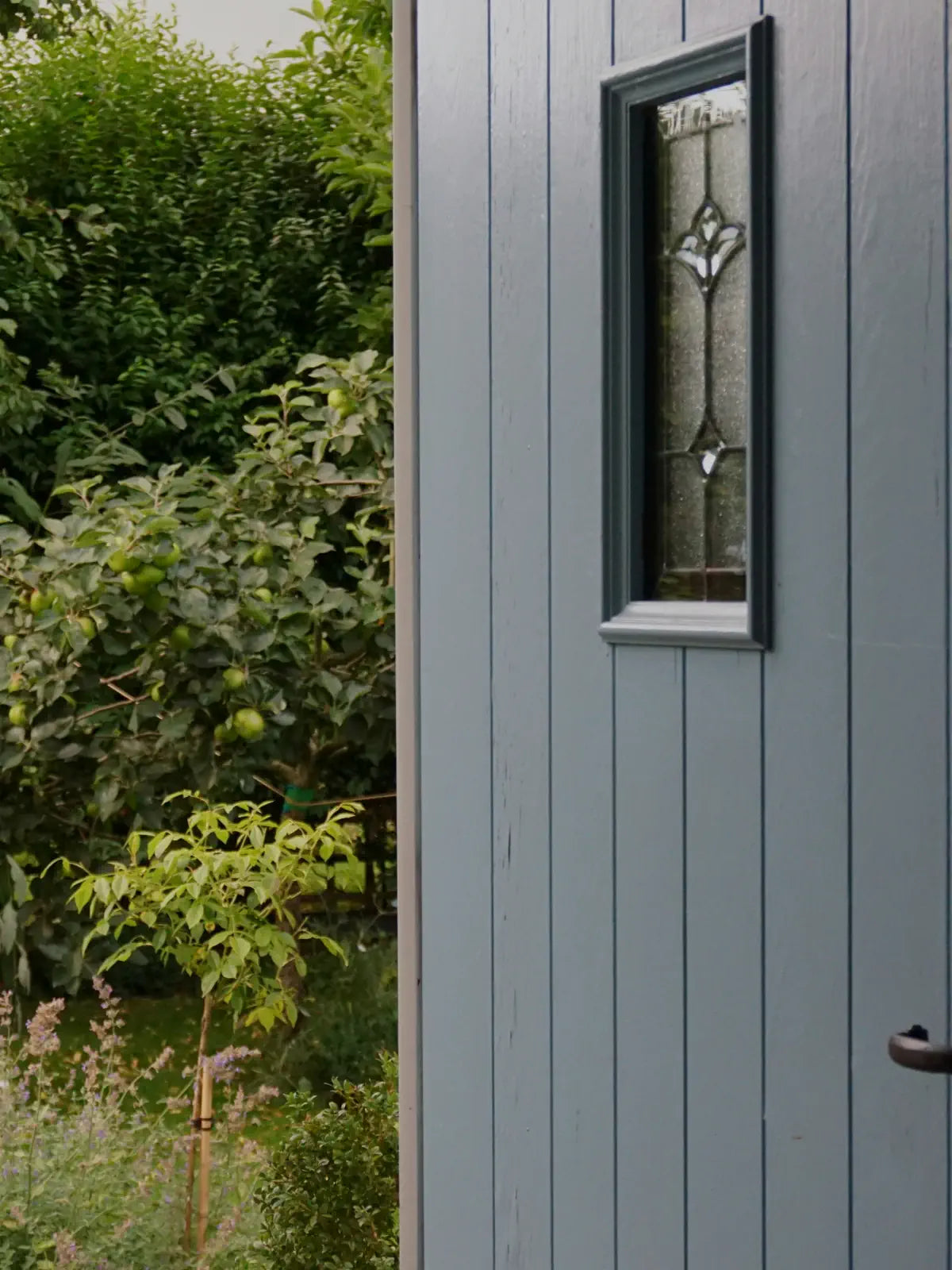 Gray wooden door with a decorative glass panel and greenery outside