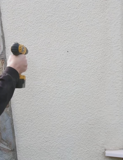 A video of a man installing a slate house sign on the front of his homes wall. He drillstwo holes in the wall with an electric drill, then installs the sign with screws using a screwdriver. At the end he gives a thumbs up to the camera.