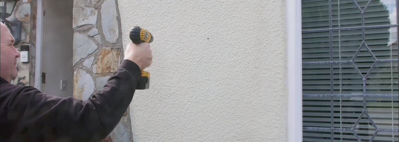 A video of a man installing a slate house sign on the front of his homes wall. He drillstwo holes in the wall with an electric drill, then installs the sign with screws using a screwdriver. At the end he gives a thumbs up to the camera.