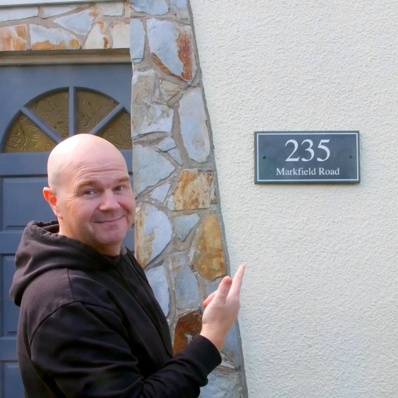 a video of a man in his living room on a white sofa talking about a grey slate sign with white engraved text on it. The sign reads 235 Markfield Road. The man shows the sign being unwrapped from brown cardboard and also being installed on a cream pebble-dashed wall.