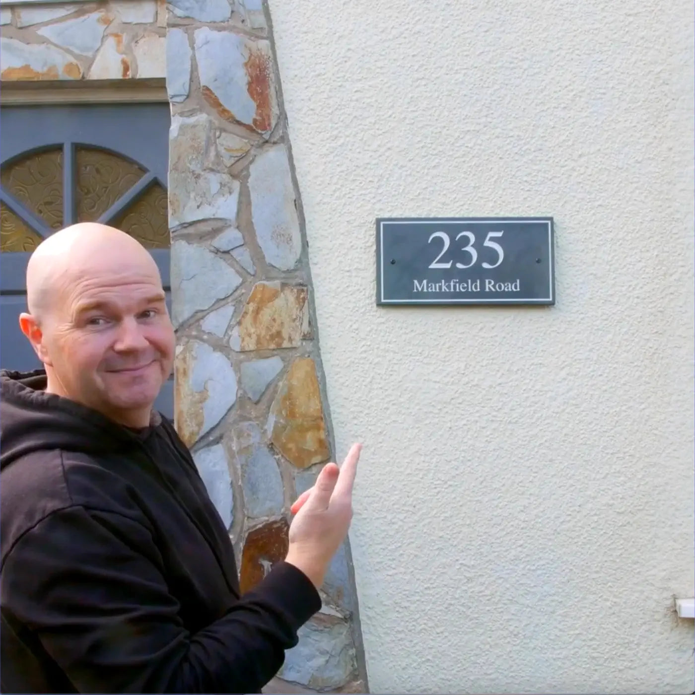 a video of a man in his living room on a white sofa talking about a grey slate sign with white engraved text on it. The sign reads 235 Markfield Road. The man shows the sign being unwrapped from brown cardboard and also being installed on a cream pebble-dashed wall.