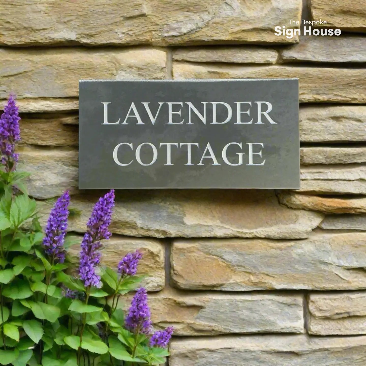 A rectangular slate house sign with the text “LAVENDER COTTAGE,” mounted on a stone wall with purple lavender flowers in the foreground.