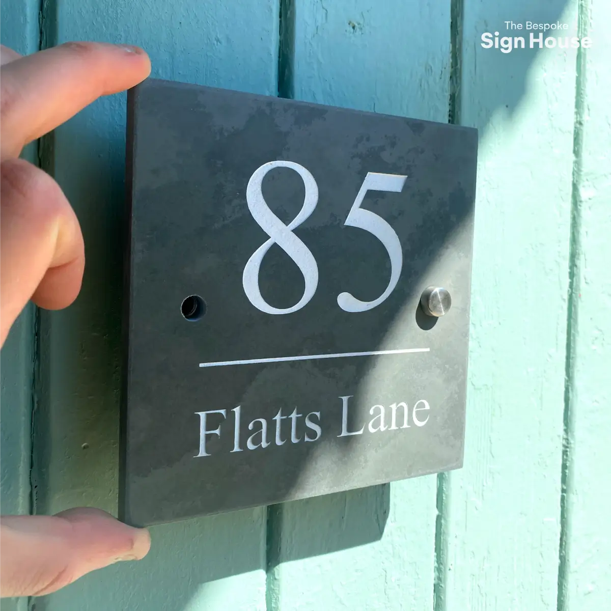 A person is installing a slate house sign that reads "85 Flatts Lane" onto a light blue wooden wall. The sign is being held in place with one hand while metal standoff caps are being screwed into pre-drilled holes. The text "The Bespoke Sign House" is displayed in the top right corner.