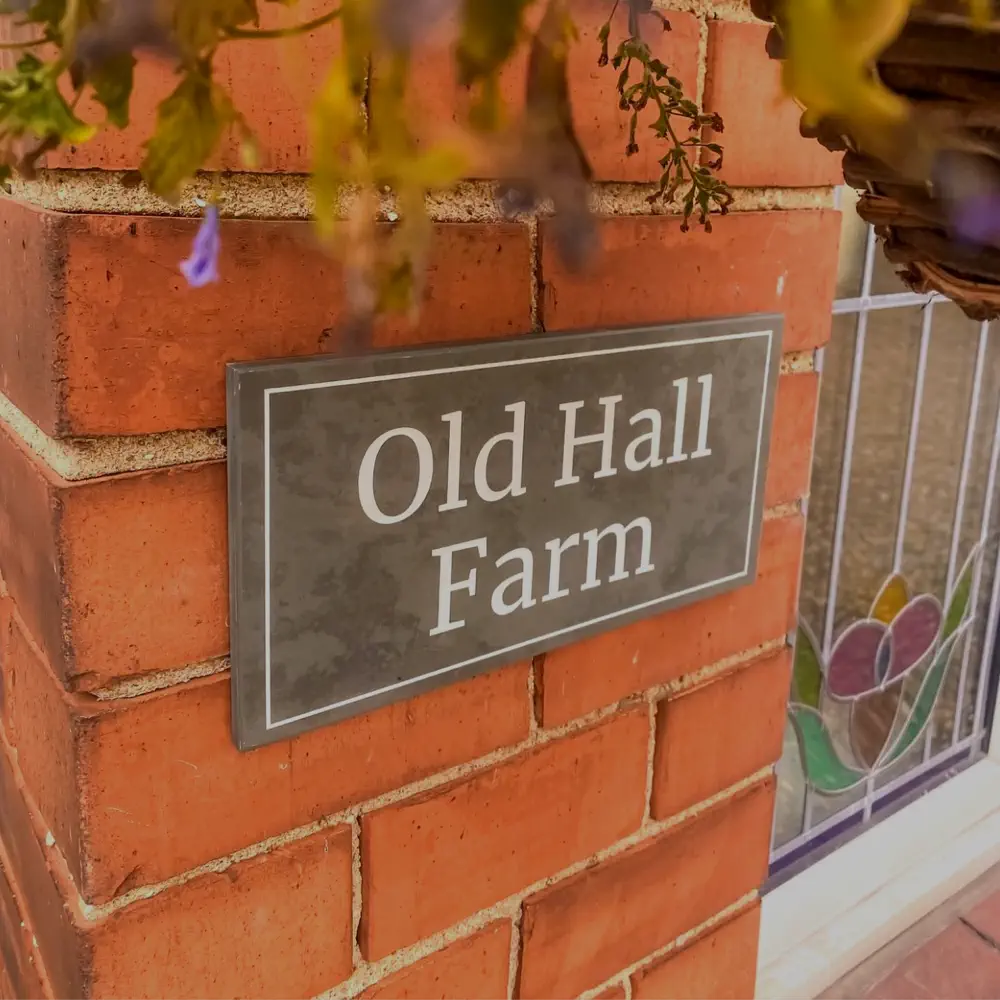 a grey slate house sign installed on a red brick wall next to window. The sign reads "Old Hall Farm"