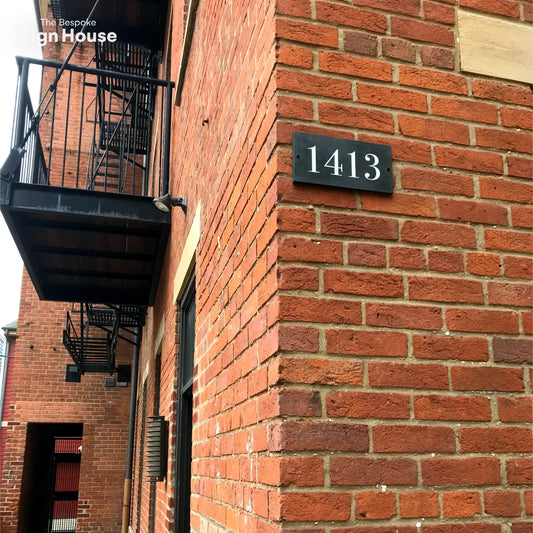  This image shows a rectangular slate sign with the number "1413" in white font mounted on a red brick wall at the corner of a building. The building features black metal fire escape stairs and balconies.