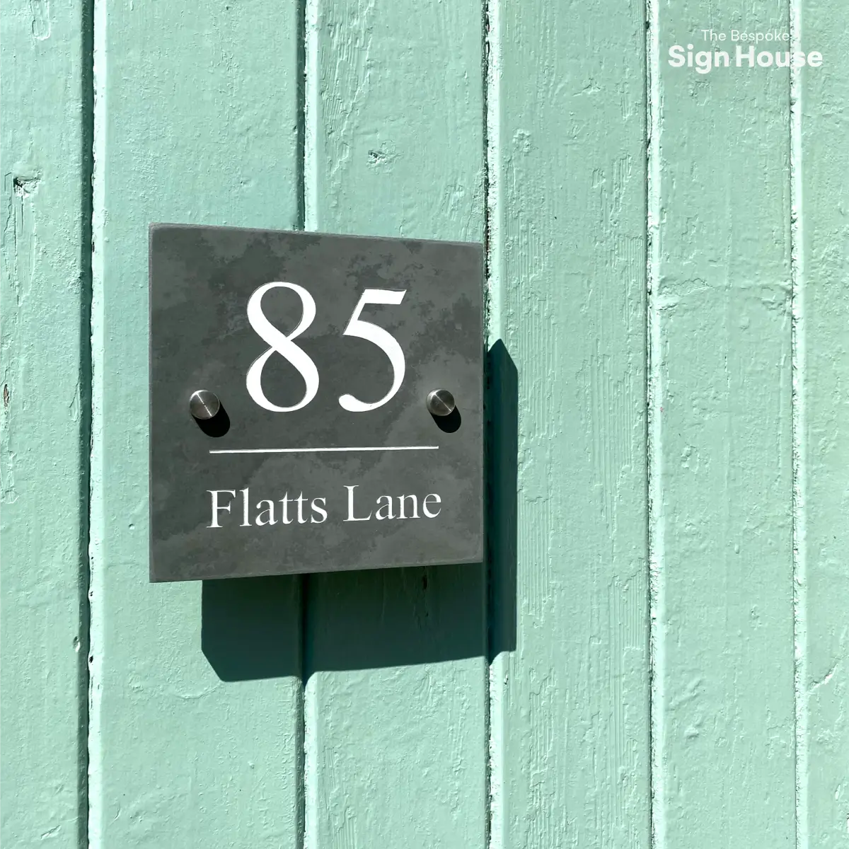 A square slate house number sign mounted on a light blue wooden wall. The sign reads "85 Flatts Lane" in white text. The sign is affixed with two stainless steel stand offs, and the shadow of the sign is visible on the wall. In the top right corner, the text "The Bespoke Sign House" is displayed.