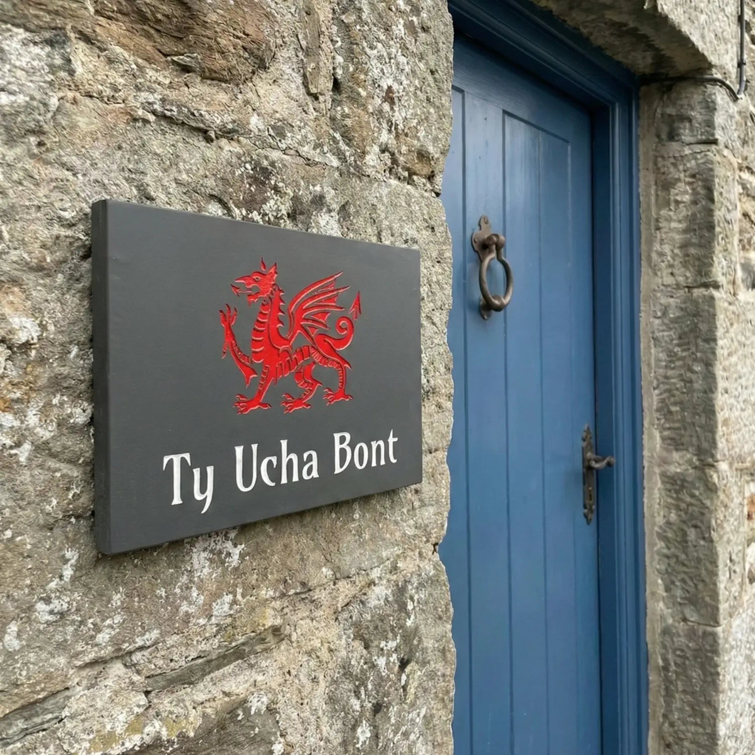 Slate house sign with red dragon emblem and text 'Ty Ucha Bont' on a stone wall.