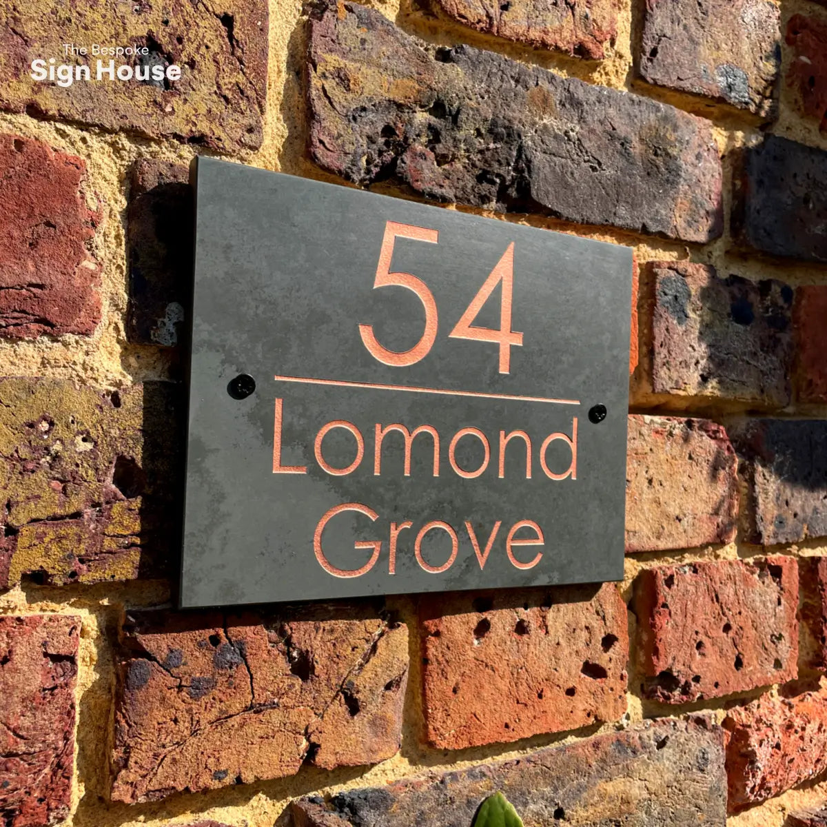 A close-up image of a slate house sign mounted on a brick wall. The sign reads “54 Lomond Grove” in modern, copper-colored lettering. The wall features rustic, red and dark bricks, with the sign attached by two small screws. The sunlight highlights the sign and the textured surface of the bricks.