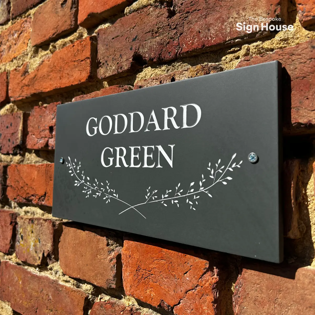 The image shows a dark slate house sign mounted on a brick wall. The sign reads “GODDARD GREEN” in white uppercase text, with a decorative design of crossed sprigs below the text. Two silver screws are visible, securing the sign to the wall. The bricks behind the sign are red and slightly weathered, giving a rustic appearance.