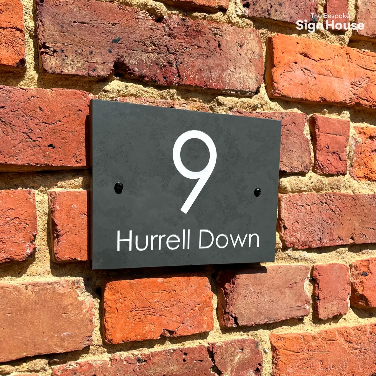 This image shows a rectangular slate sign mounted on a red brick wall. The sign displays the number "9" in large white font, centered above the text "Hurrell Down" in smaller white font. The sign has two small black screws on either side securing it to the wall.