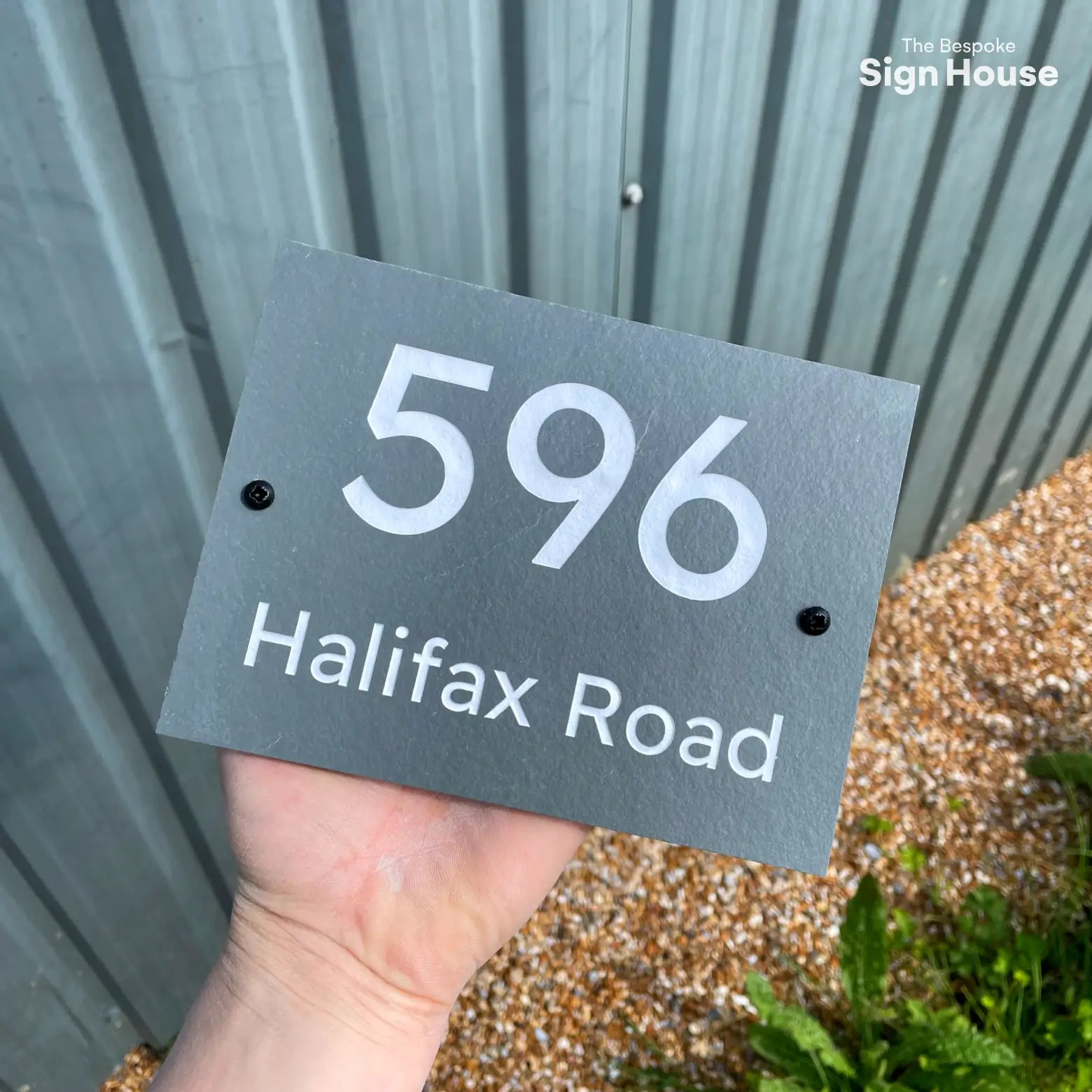 This image shows a hand holding a rectangular slate sign with the number "596" in large white font, centered above the text "Halifax Road" in smaller white font. The background includes a grey corrugated metal wall and a gravel ground with some green plants. There are two small black screws on either side of the sign.