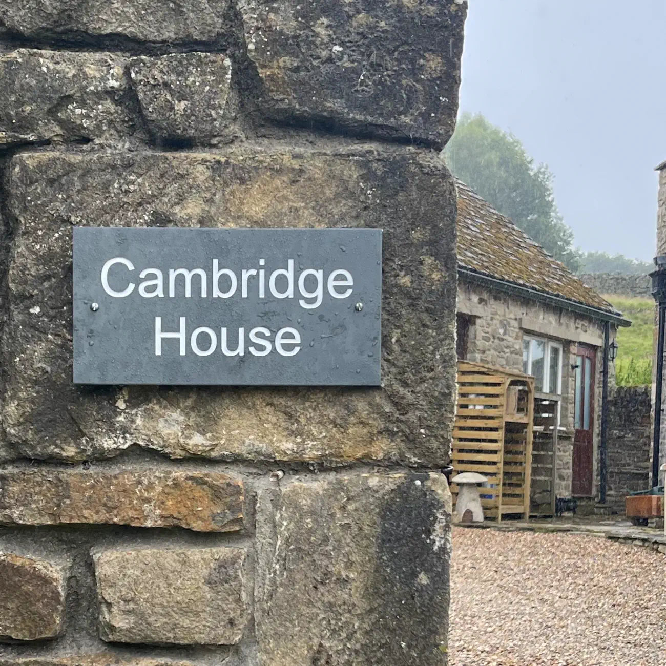 Grey slate sign with 'Cambridge House' on a stone wall next to a farm entrance