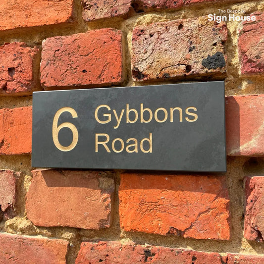 a smooth grey slate house sign with a gold number 6 and gybbons road next to it on a red brick wall