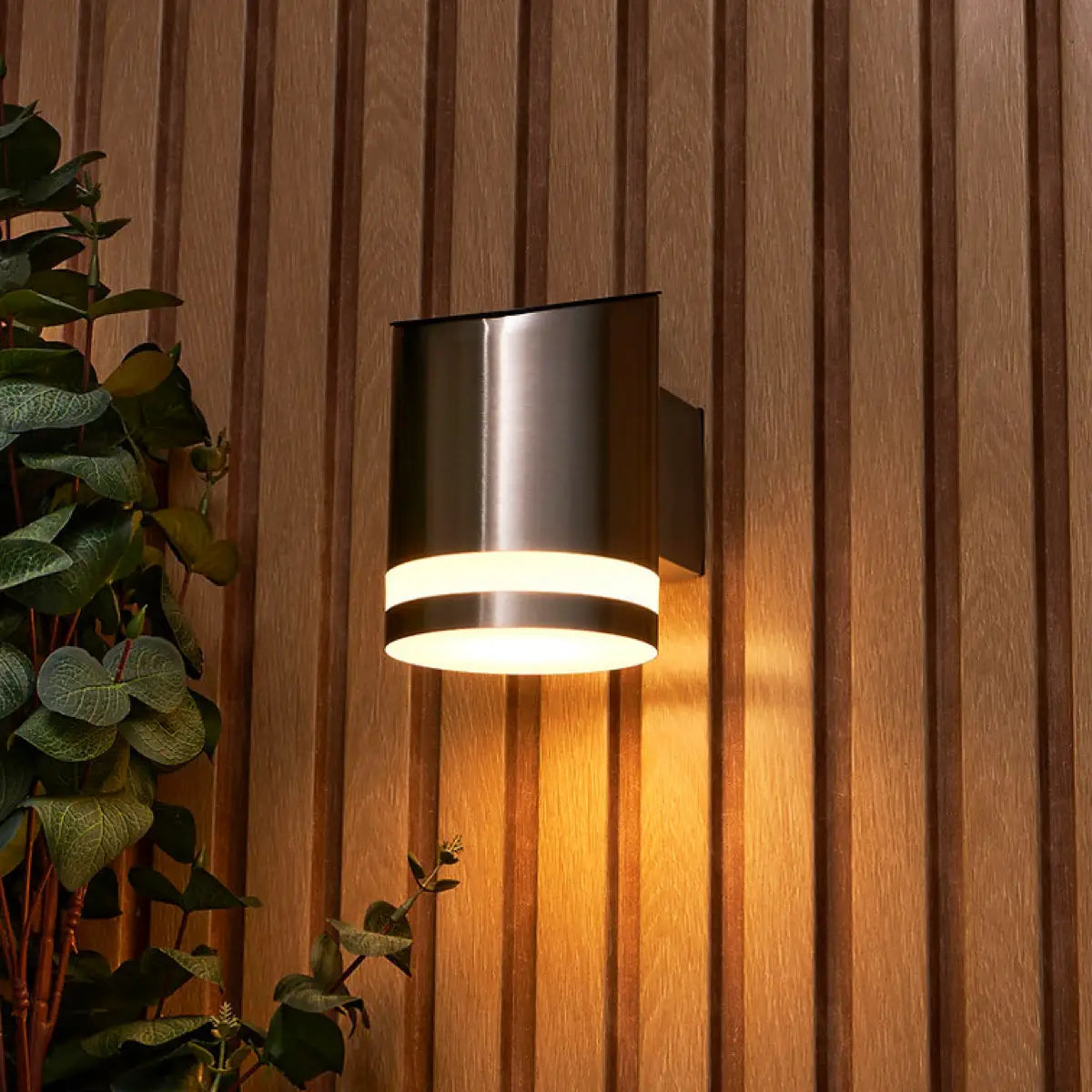 A sleek, modern, cylindrical solar-powered outdoor wall light with a brushed metal finish, emitting a warm glow. The light is mounted on a vertical wooden panel wall with a striped pattern. To the left of the light, there is a green plant with round leaves, adding a natural element to the scene. The light fixture casts a soft, inviting light, enhancing the warmth and texture of the wooden wall.
