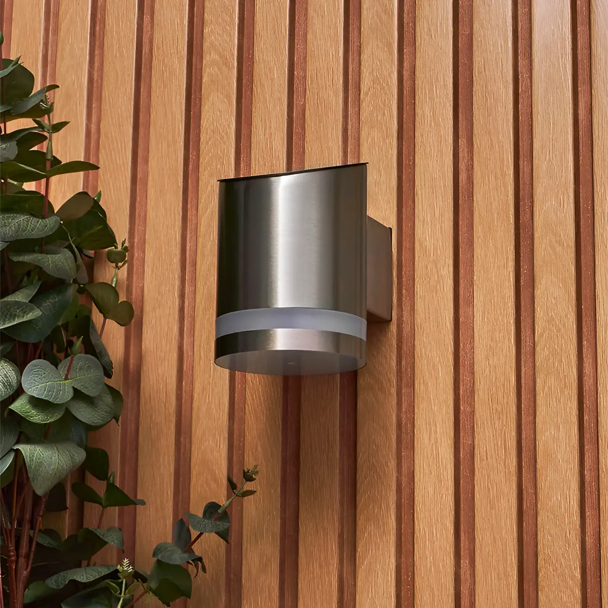 A sleek, modern, cylindrical solar-powered outdoor wall light with a brushed metal finish, mounted on a vertical wooden panel wall with a striped pattern. To the left of the light, there is a green plant with round leaves, adding a touch of natural greenery to the scene. The light fixture has a contemporary design that complements the wooden texture of the wall.