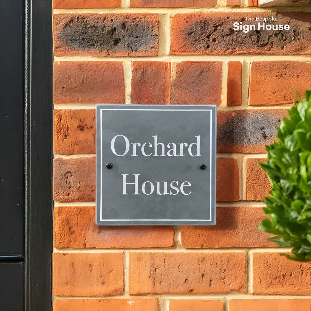 A square slate house sign with white lettering that reads “Orchard House,” mounted on a red brick wall next to a black door frame. A neatly trimmed green shrub is visible in the foreground.