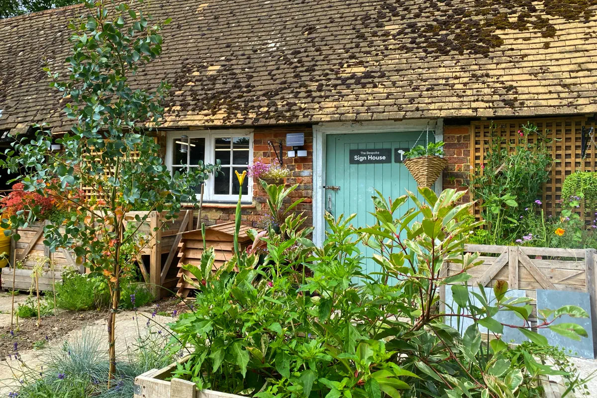 The image shows a charming brick workshop with a moss-covered roof and a pale green door labeled “The Bespoke Sign House.” The front garden is lush with various plants and flowers, adding a vibrant touch to the scene. A hanging basket and wooden structures are visible in the background.