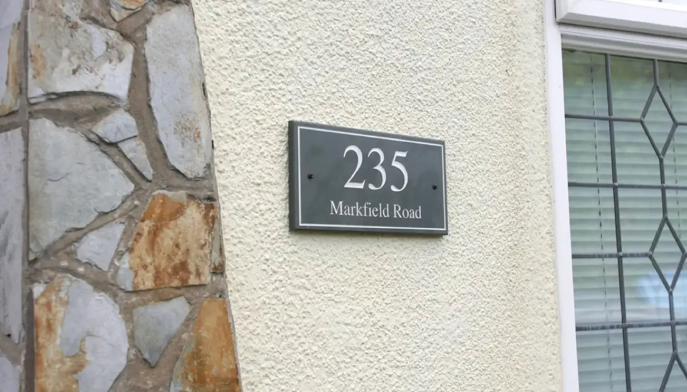 This is an image showing a modern house sign with the text “235 Markfield Road” mounted on a wall with a mix of textured plaster and stone elements.