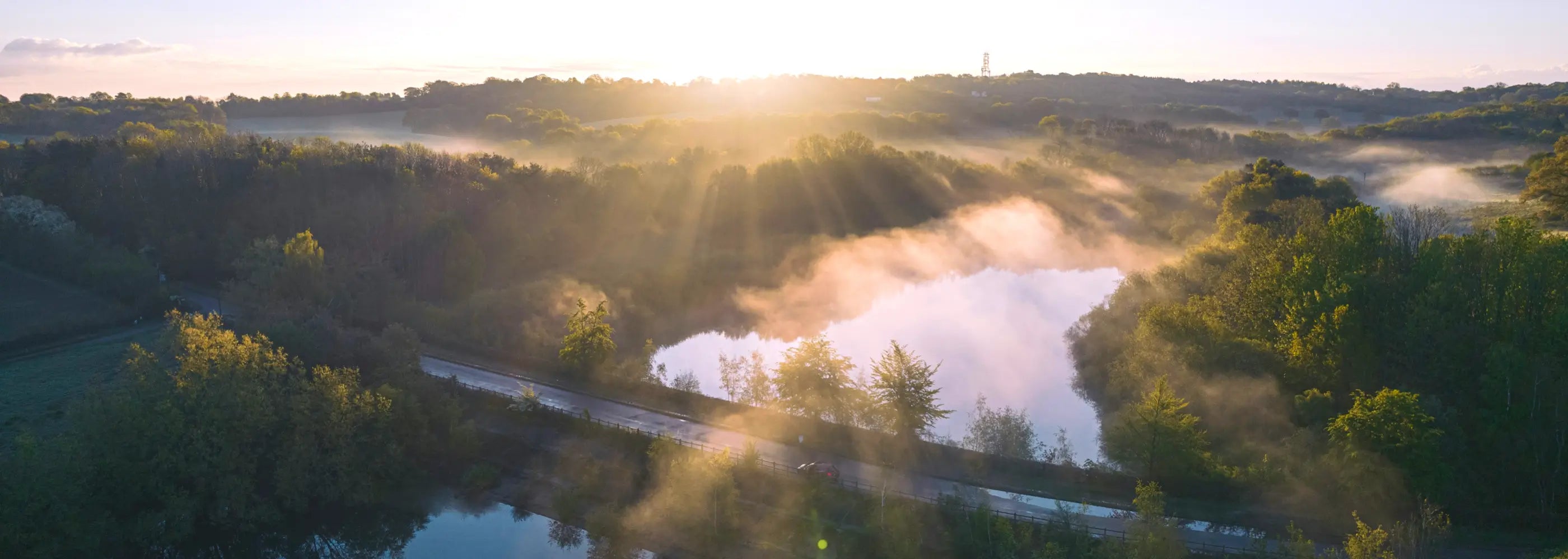 A breathtaking sunrise over a misty forested landscape, with sunlight streaming through the trees and reflecting off a tranquil lake. The scene exudes a peaceful, ethereal quality with its soft morning light and mist.