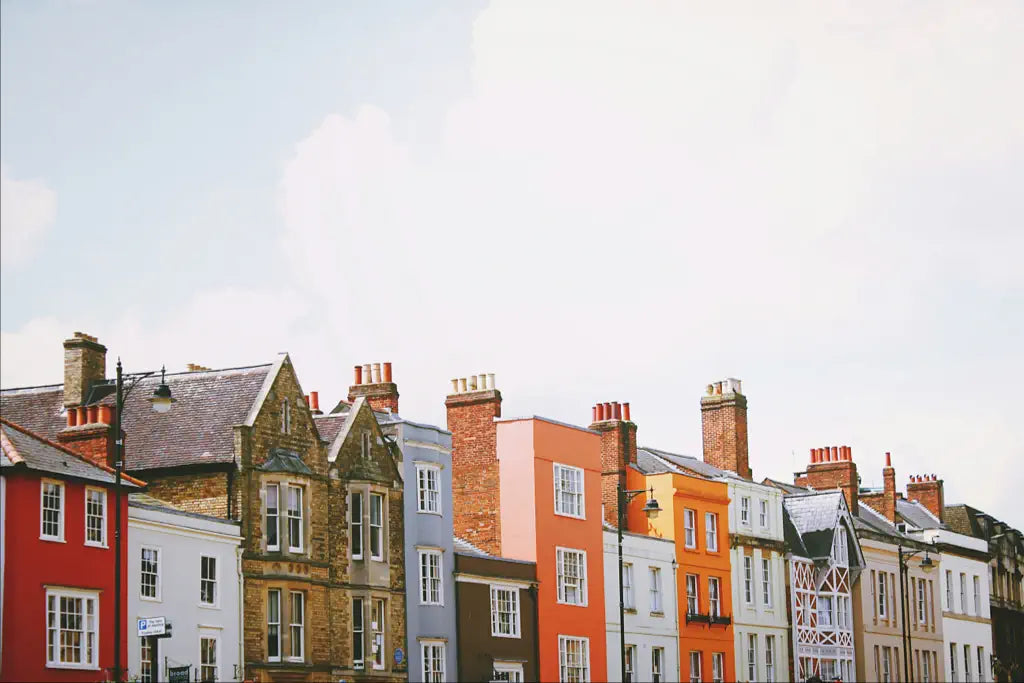 A row of colorful, historic terraced houses with chimneys on a clear day. The buildings vary in architectural style and color, ranging from red and white to grey and orange, against a bright sky.