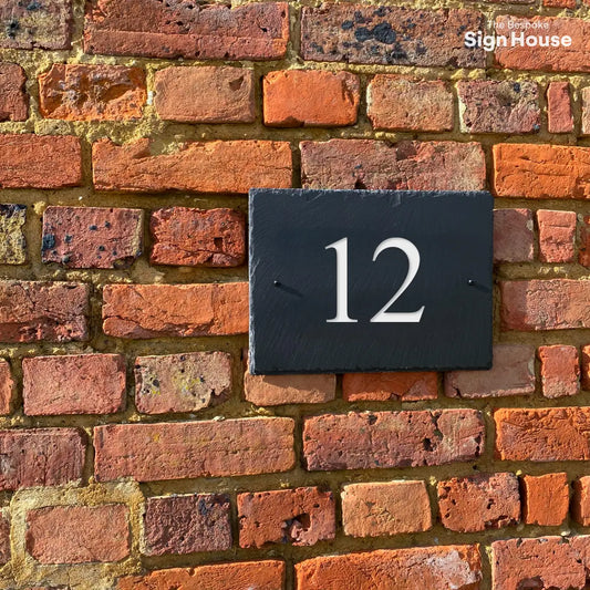 a black sign made using welsh slate. The sign is screwed into a red brick wall with a number 12 engraved onto it and painted white. The font is times new roman 